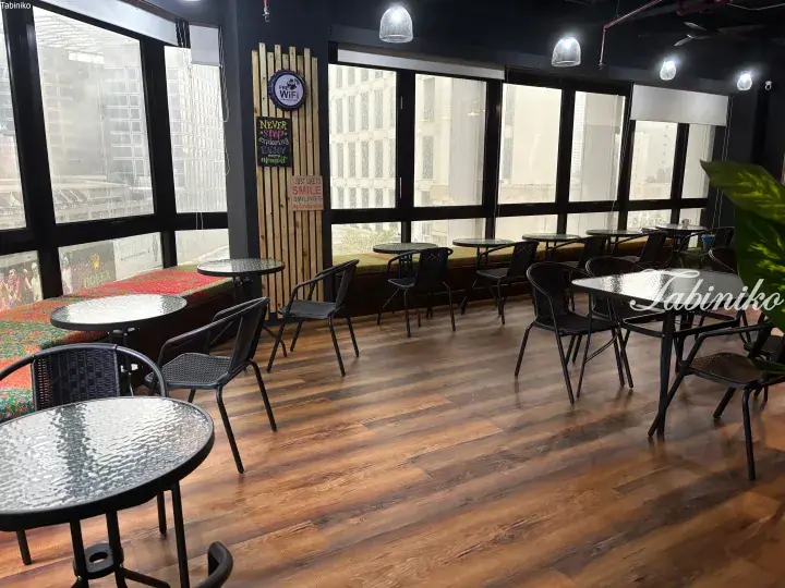 Modern cafe interior with wooden floors, black wicker chairs, and large windows overlooking a city view.