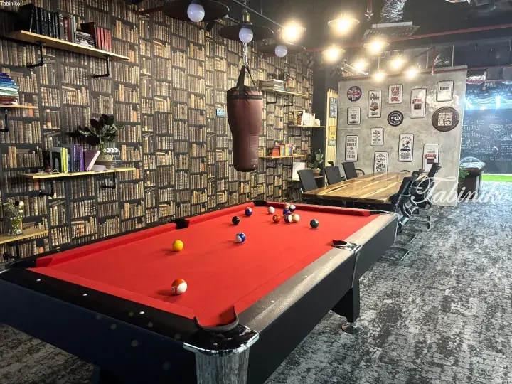 Red pool table in a stylish lounge with bookshelf wallpaper, hanging punching bag, and wooden meeting table.