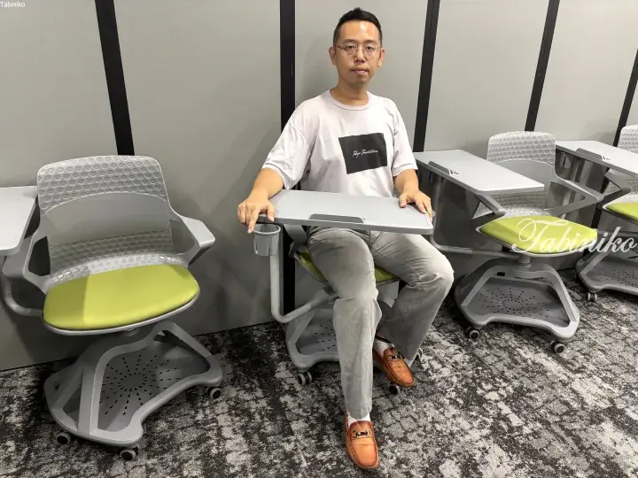 Man demonstrating a modern grey and green study chair with an attached desk in a showroom.