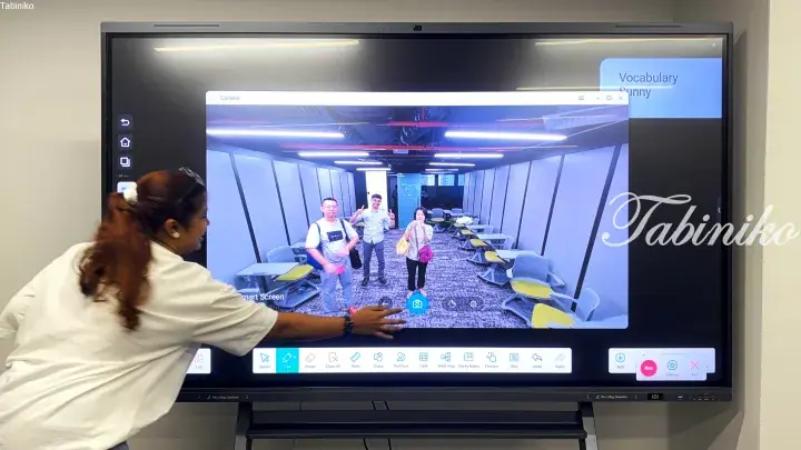 Woman interacting with a large smart screen displaying a camera view of a modern co-working space with yellow chairs.