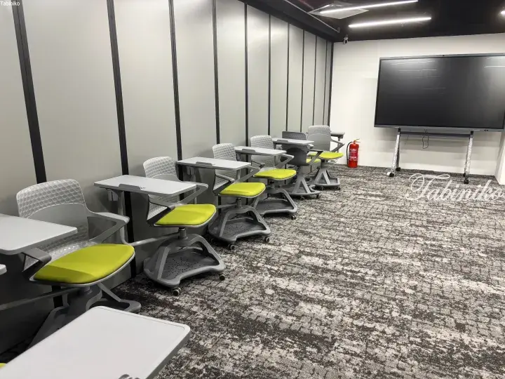 Modern classroom with adjustable green chairs and white desks arranged in rows facing a large digital screen.