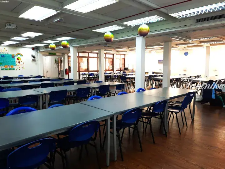 Spacious school canteen with long grey tables, blue chairs, and festive balloons hanging from the ceiling.
