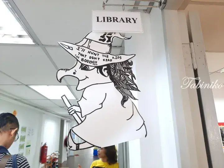 Whimsical witch cutout sign at a library entrance with text encouraging kids to read books.