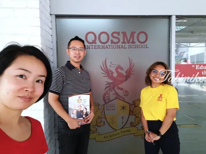 Three people standing in front of QOSMO International School’s glass entrance featuring a phoenix crest.