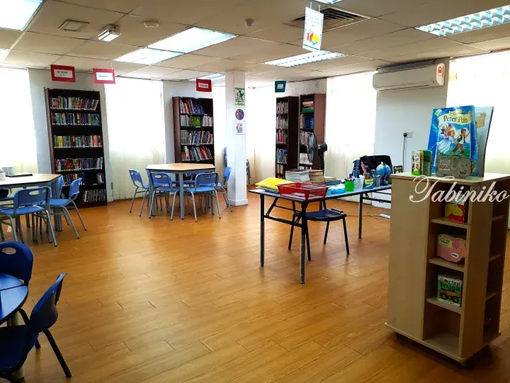 Bright school library with wooden floors, bookshelves, and blue chairs arranged around tables.