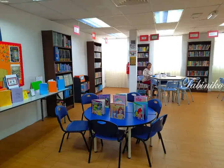 Bright school library with blue children’s reading table, bookshelves, and a teacher at a desk.