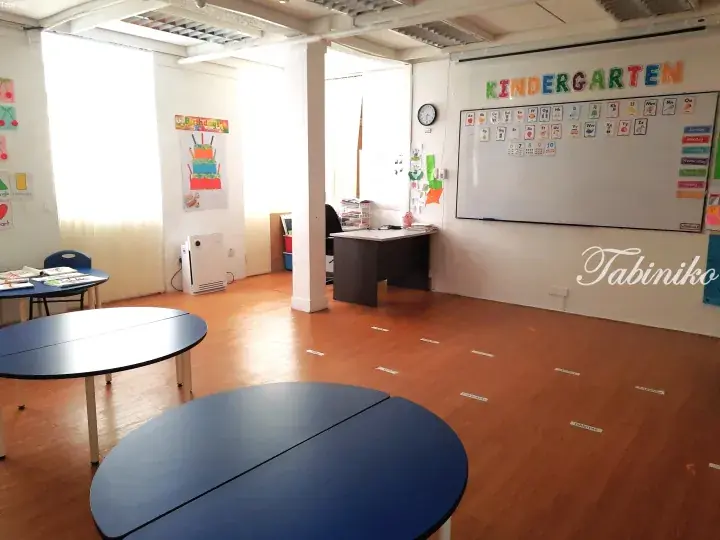 Bright kindergarten classroom with blue tables, alphabet posters, and name tags on the wooden floor.
