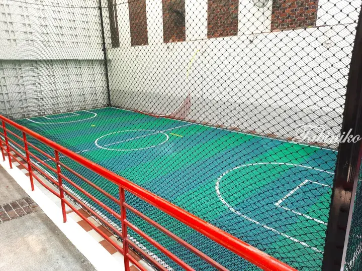 Green and blue basketball court with white lines enclosed by a black safety net and red railing.