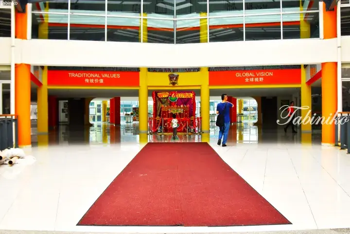 A festive Deepavali display with a red carpet leads through a modern building lobby featuring pillars and signs for Traditional Values and Global Vision.