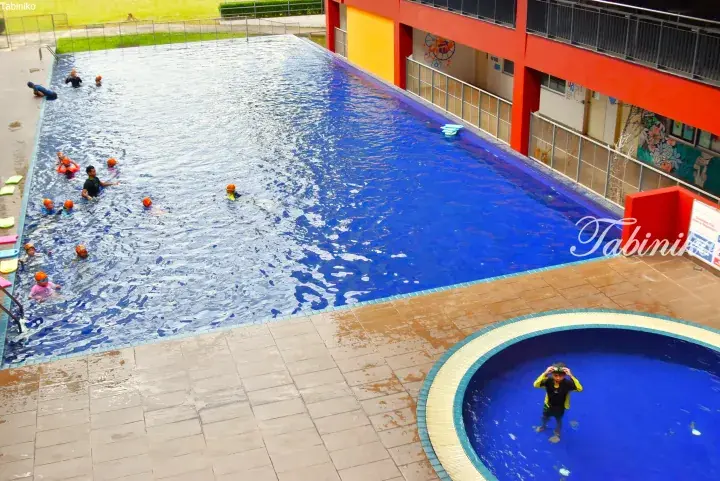 Children in orange caps learn swimming in a blue pool at a Malaysia school, with a child in a separate round pool.