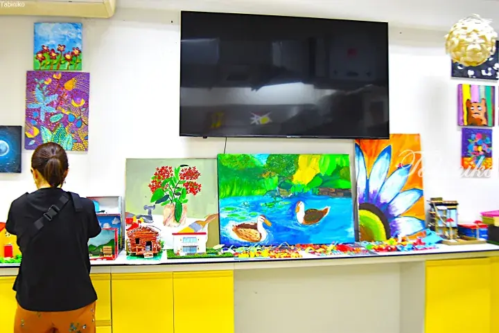 Woman viewing children’s colorful paintings and crafts displayed on a counter in a bright art studio.