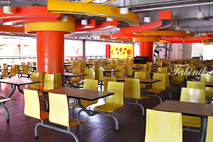 Bright cafeteria with yellow chairs, orange pillars, and swirling ceiling lights.