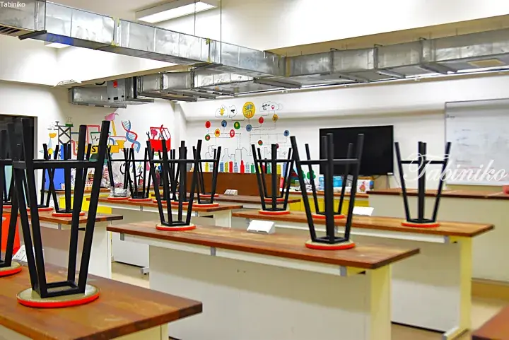 Classroom with chairs stacked upside down on wooden desks, featuring exposed ceiling ducts and colorful wall murals.