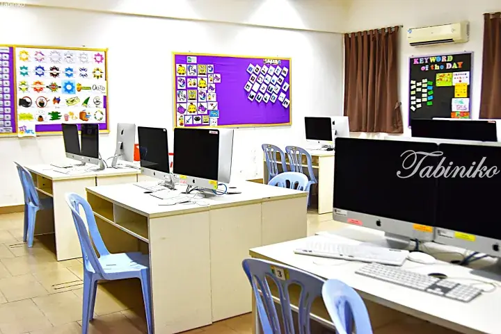 Bright computer lab with Apple iMacs, blue chairs, and colorful educational bulletin boards.