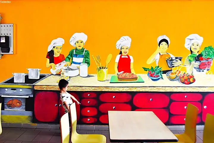 Child looking at a vibrant kitchen mural depicting chefs cooking, likely inside a Malaysian family-friendly restaurant.