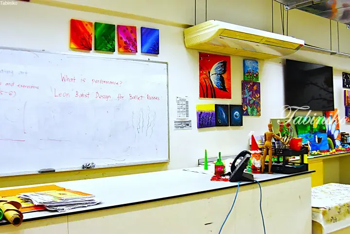 Art classroom with whiteboard showing ‘Leon Bakst Design for Ballet Russes’ and colorful student paintings on walls.