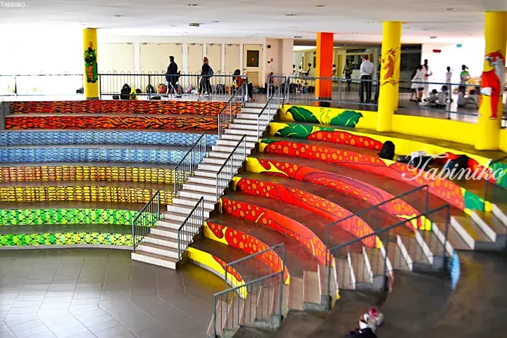 Colorful patterned stairs inside a modern building with yellow pillars and people walking on the upper level.