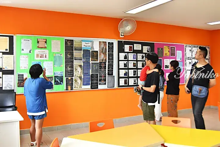 Visitors viewing student poetry and playscript displays on a green bulletin board in a bright orange-walled classroom.
