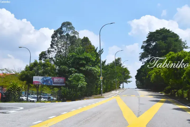 A quiet road with yellow markings leads uphill past lush green trees and a banner for REAL Schools in Malaysia.