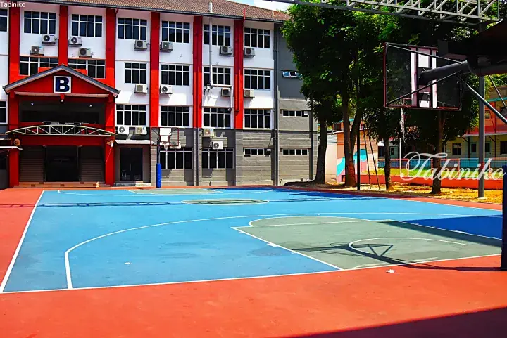 Outdoor basketball court with blue and red flooring in front of a school building marked with a large ‘B’.