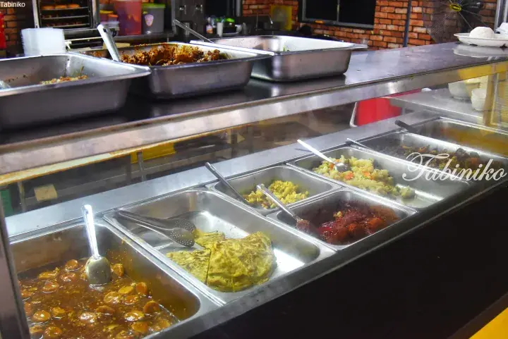 Variety of Malaysian dishes displayed in stainless steel trays at a food stall counter.