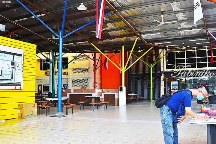 A man signs in at a school registration desk in a colorful open-air Malaysian campus with flags and seating areas.