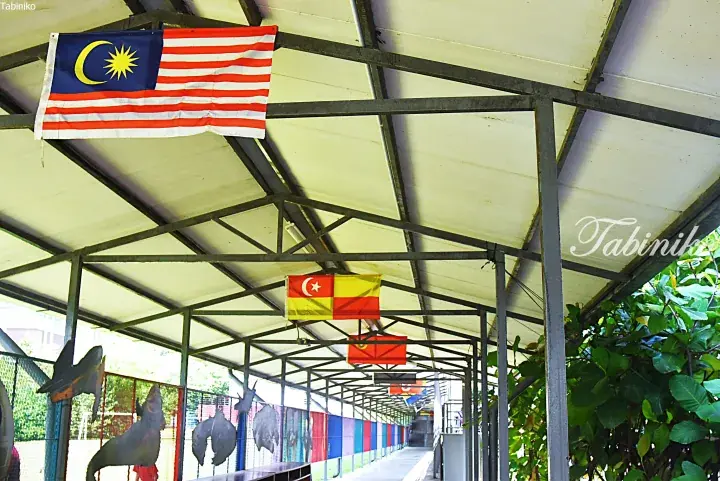 Malaysia’s national flag and state flags hang from a covered walkway roof, with colorful metal animal silhouettes along the fence.