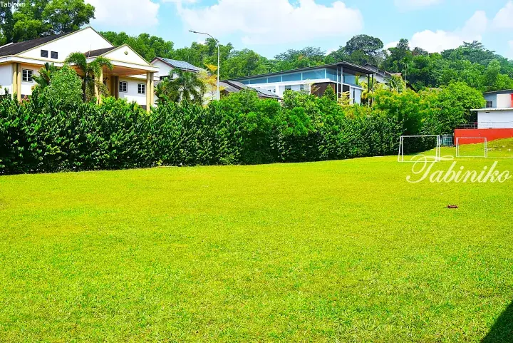 Lush green lawn with soccer goals in front of a modern house and dense hedge in a Malaysian residential area.