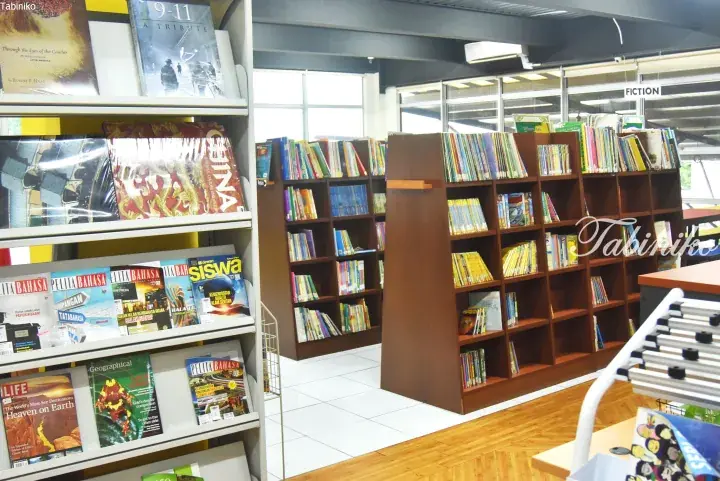 Interior view of a library with bookshelves displaying magazines and fiction books in a well-lit space.