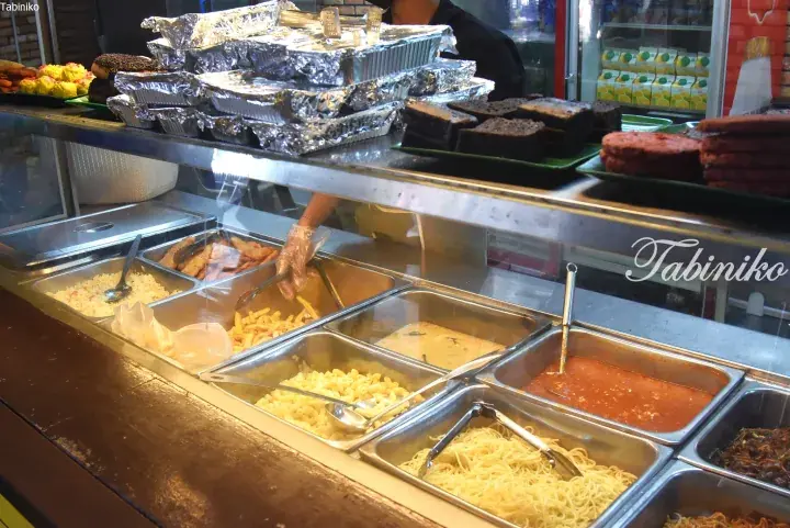 A food stall display featuring various Malaysian dishes like noodles, curry, and fried items in stainless steel containers.