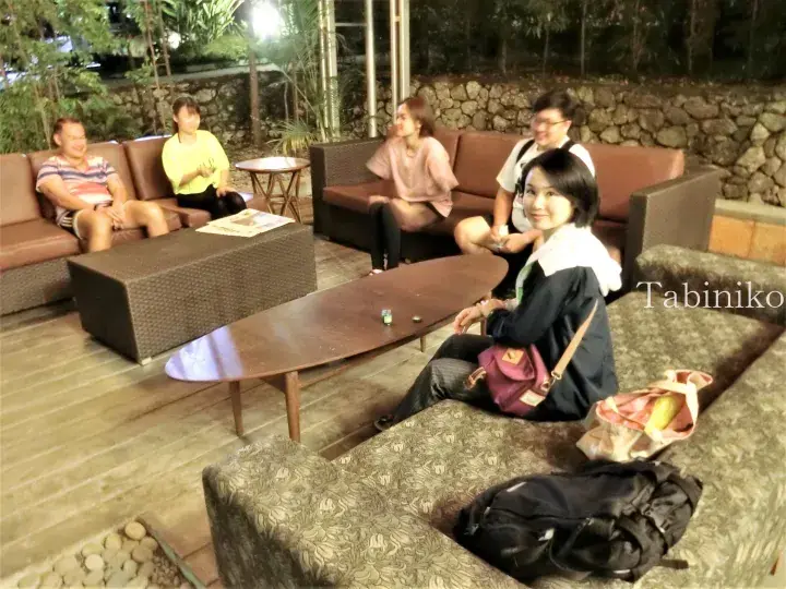 Group of friends relaxing on outdoor sofas at night in a tropical resort setting with wooden decking and lush greenery.