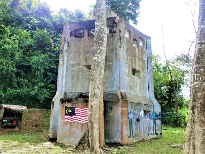 Concrete bunker with Malaysian flag and mural of historical figures in a lush forest setting.