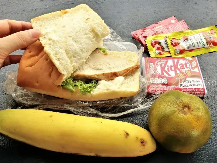 A hand lifts the top of a chicken sandwich revealing lettuce and mayo, alongside a banana, mango, KitKat bar, and ketchup sachets.