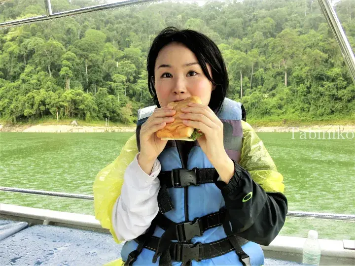 Woman in life jacket eating a sandwich on a boat with green water and forested hills in the background.