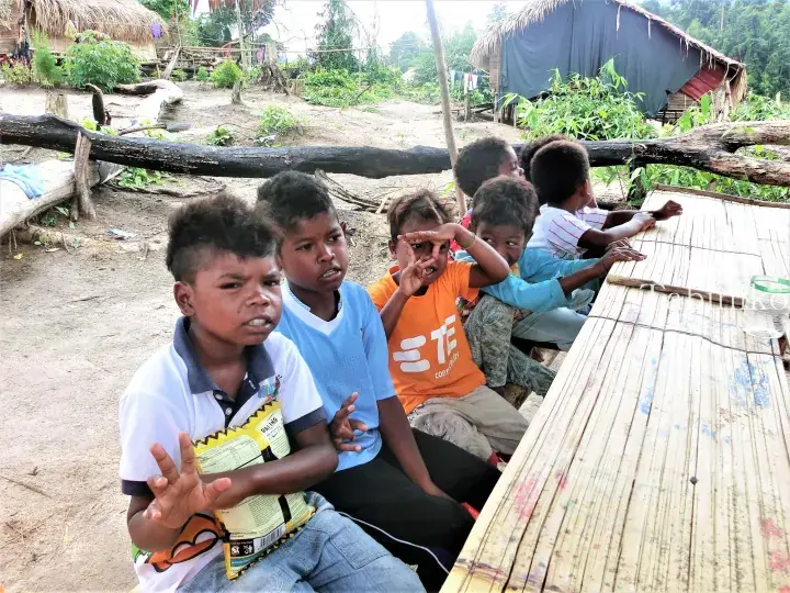 Children sitting at a bamboo table in a rural village setting with traditional thatched huts in the background.