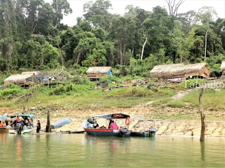 Tour boats docked at a riverbank near traditional thatched-roof huts in a lush Malaysian jungle setting.