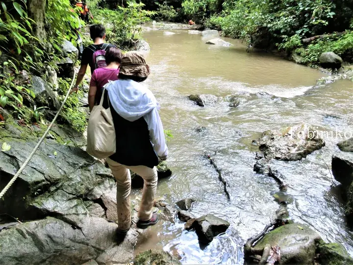 Hikers navigate rocky terrain beside a stream in a lush Malaysian jungle.