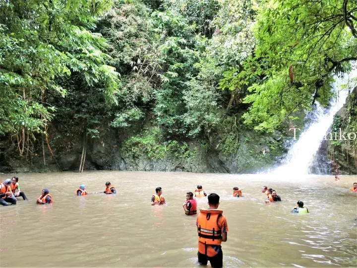 Tourists wearing life jackets swim in a river pool at the base of a waterfall in a lush Malaysian jungle.