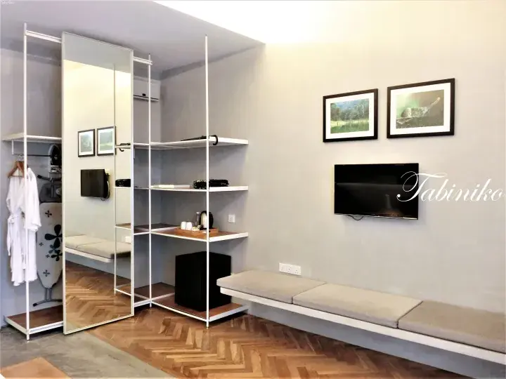 Modern hotel room interior with a white shelving unit, mirrored wardrobe, and a long cushioned bench under a wall-mounted TV.