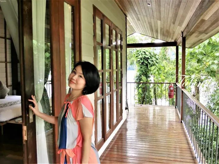 Woman standing on a wooden balcony at a resort, opening a glass door with lush greenery and water visible outside.