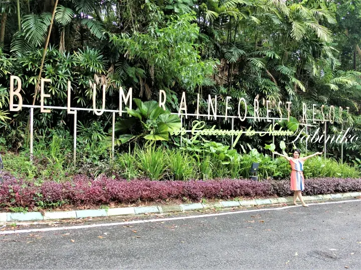 Woman posing at the entrance sign of Belum Rainforest Resort in Malaysia, surrounded by lush tropical greenery.