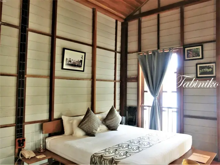 Cozy bedroom with wooden walls, a neatly made bed, and a window with blue curtains overlooking a balcony.