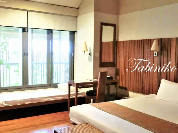 Bright hotel room with wooden accents, a bed, desk, and large window overlooking greenery.