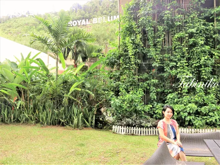 Woman sitting on a woven bench at Royal Belum Eco Resort, surrounded by lush tropical greenery and a hillside sign.