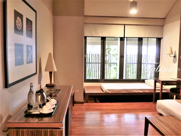 Spacious hotel room with wooden floors, a desk with tea set, and a daybed by the window overlooking greenery.