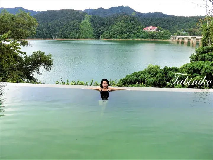 Woman relaxing in an infinity pool overlooking a serene lake and lush green hills in Malaysia.