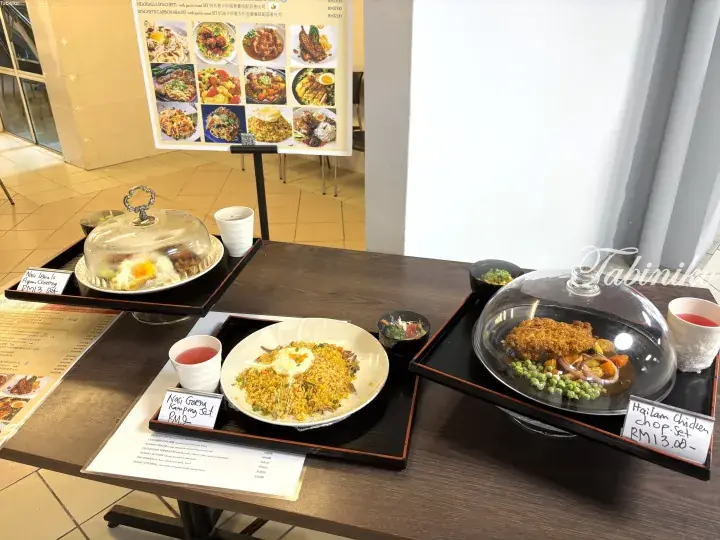 Three Malaysian set meals displayed on wooden trays with glass domes, featuring Nasi Goreng Kampung, Hailam Chicken Chop, and Nasi Lemak Ayam Goreng.