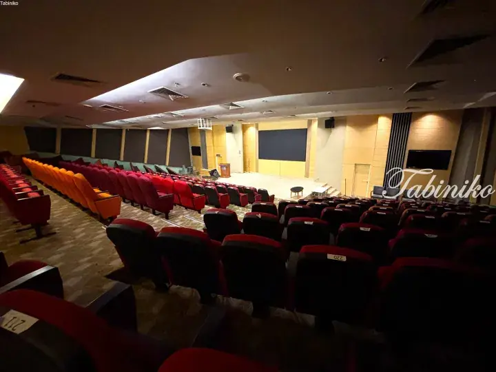 Interior view of a modern auditorium with tiered seating, red and orange chairs, and a stage with a large screen.