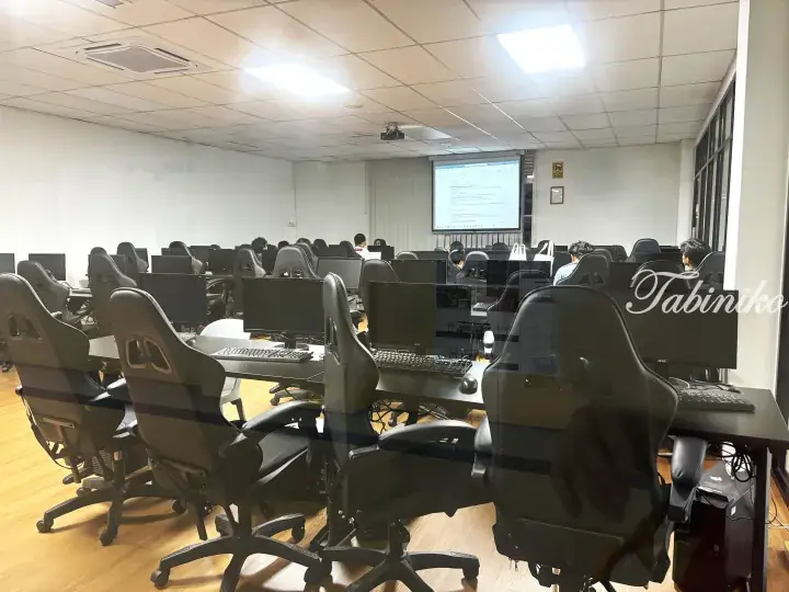 Rows of black gaming chairs and computers in a modern training room with a projector screen.