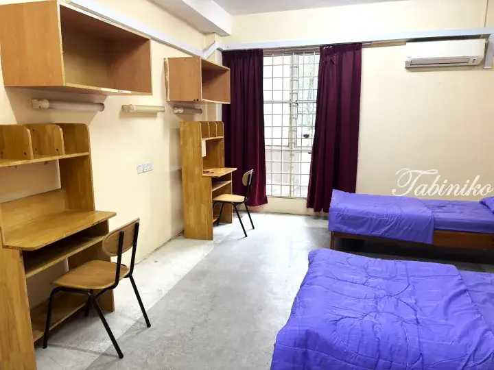 Spacious student dorm room with wooden study desks, purple bedding, and air conditioning.